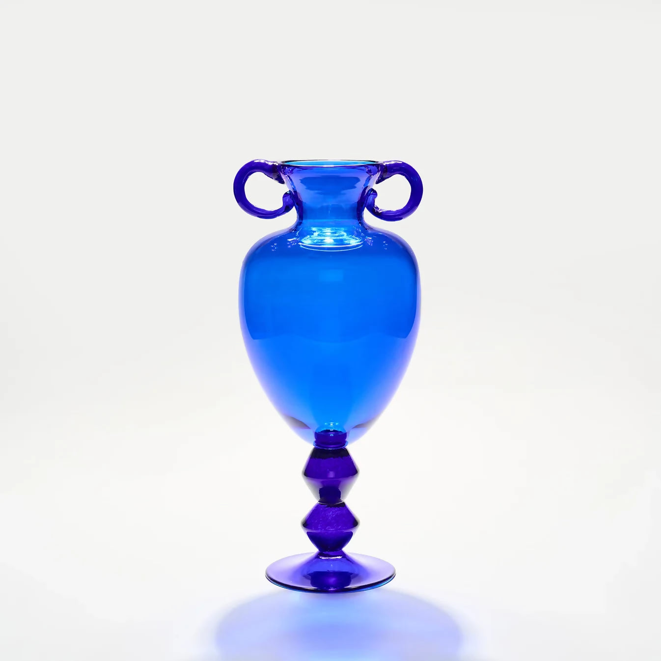Blue glass vase with handles on a white background,  available  to buy on the-salon.com. SALON curates the finest , unique, handmade pieces from the interior industry's best makers and brands. Shop luxury furniture, lighting, rugs, art, antiques and vintage pieces and accessories from SALON interior designed rooms.