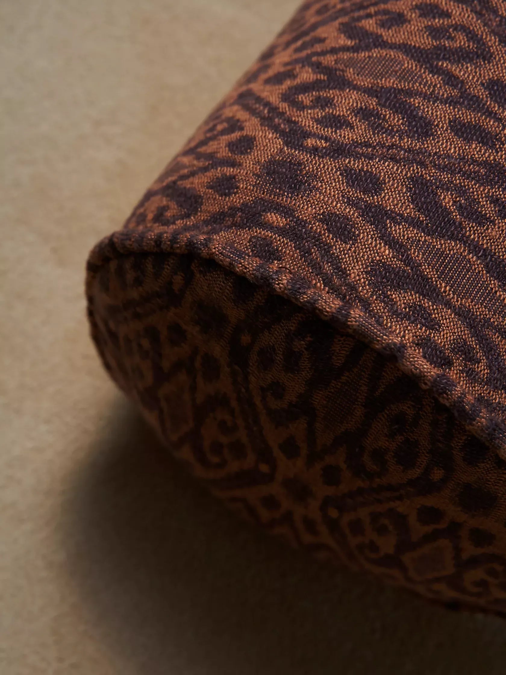 A close up of a brown cylindrical pillow with a leopard print pattern on a beige background,  available  to buy on the-salon.com. SALON curates the finest , unique, handmade pieces from the interior industry's best makers and brands. Shop luxury furniture, lighting, rugs, art, antiques and vintage pieces and accessories from SALON interior designed rooms.