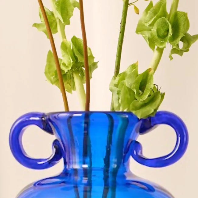 Blue glass vase with handles containing green plants on a light background,  available  to buy on the-salon.com. SALON curates the finest , unique, handmade pieces from the interior industry's best makers and brands. Shop luxury furniture, lighting, rugs, art, antiques and vintage pieces and accessories from SALON interior designed rooms.