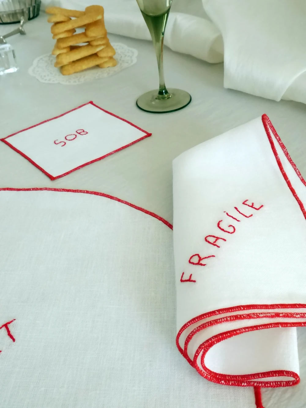 White tablecloth with red border on a white surface featuring 'fragile' hand sewn on in red  available  to buy on the-salon.com. SALON curates the finest , unique, handmade pieces from the interior industry's best makers and brands. Shop luxury furniture, lighting, rugs, art, antiques and vintage pieces and accessories from SALON interior designed rooms.