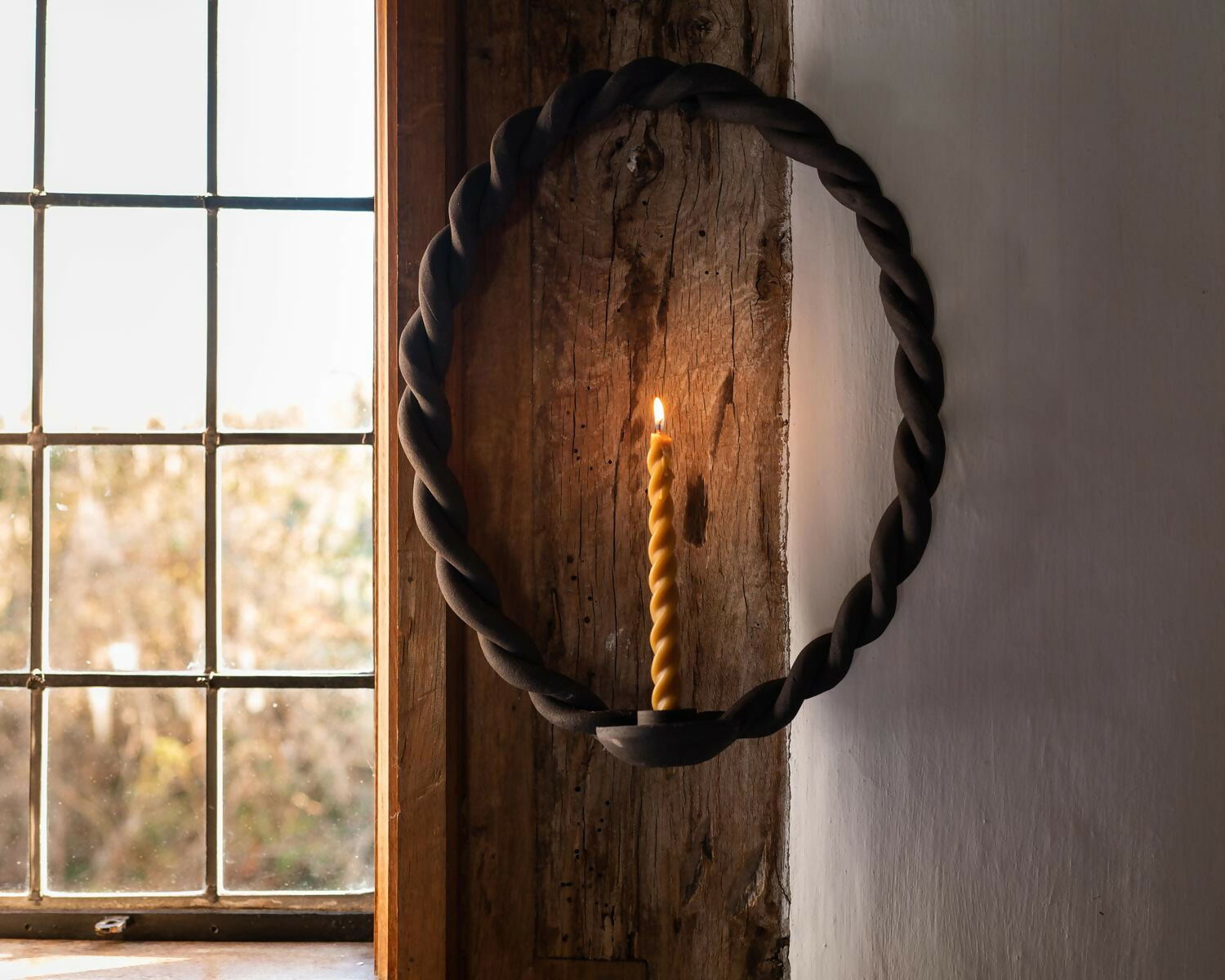 Rope candle holder with a lit candle against a wooden wall and window,  available  to buy on the-salon.com. SALON curates the finest , unique, handmade pieces from the interior industry's best makers and brands. Shop luxury furniture, lighting, rugs, art, antiques and vintage pieces and accessories from SALON interior designed rooms.
