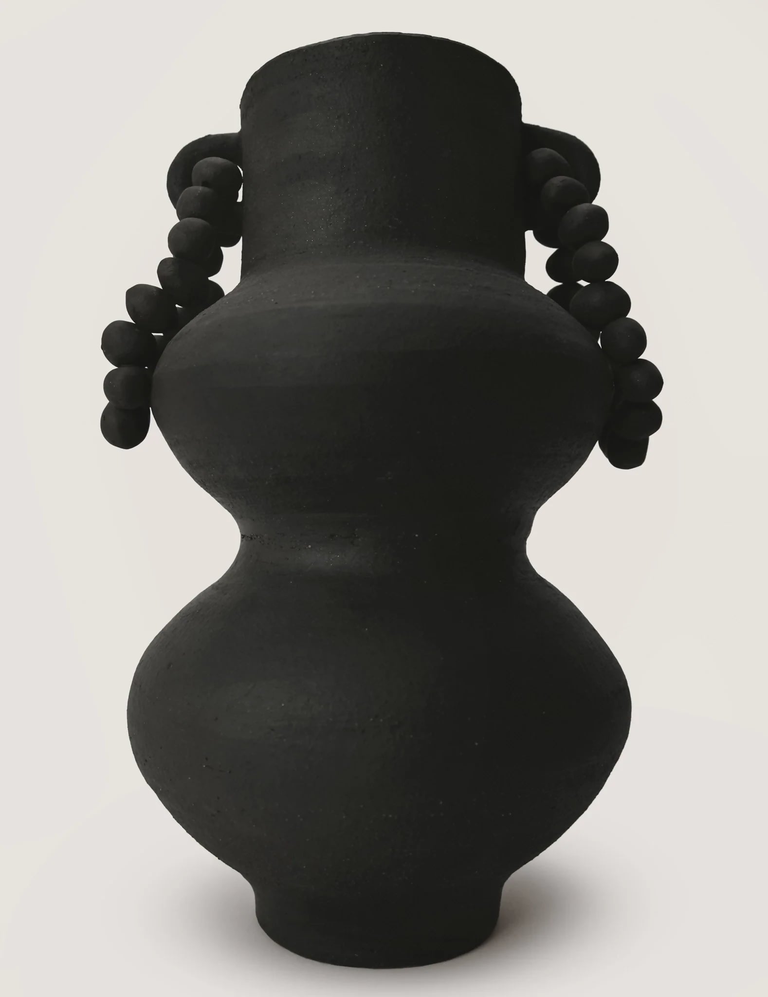 Black ceramic vase with braid-like handles on a white background,  available  to buy on the-salon.com. SALON curates the finest , unique, handmade pieces from the interior industry's best makers and brands. Shop luxury furniture, lighting, rugs, art, antiques and vintage pieces and accessories from SALON interior designed rooms.